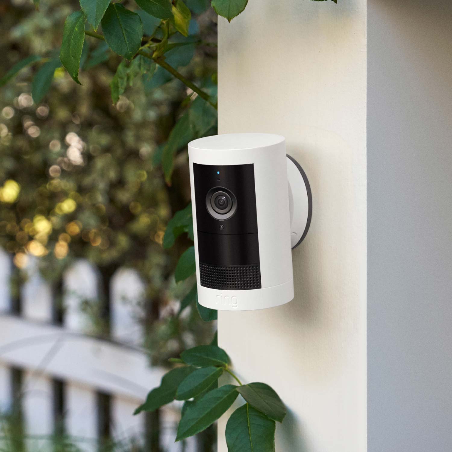 White Ring outdoor security camera mounted on an outdoor post near green leaves and a white picket fence in soft daylight.