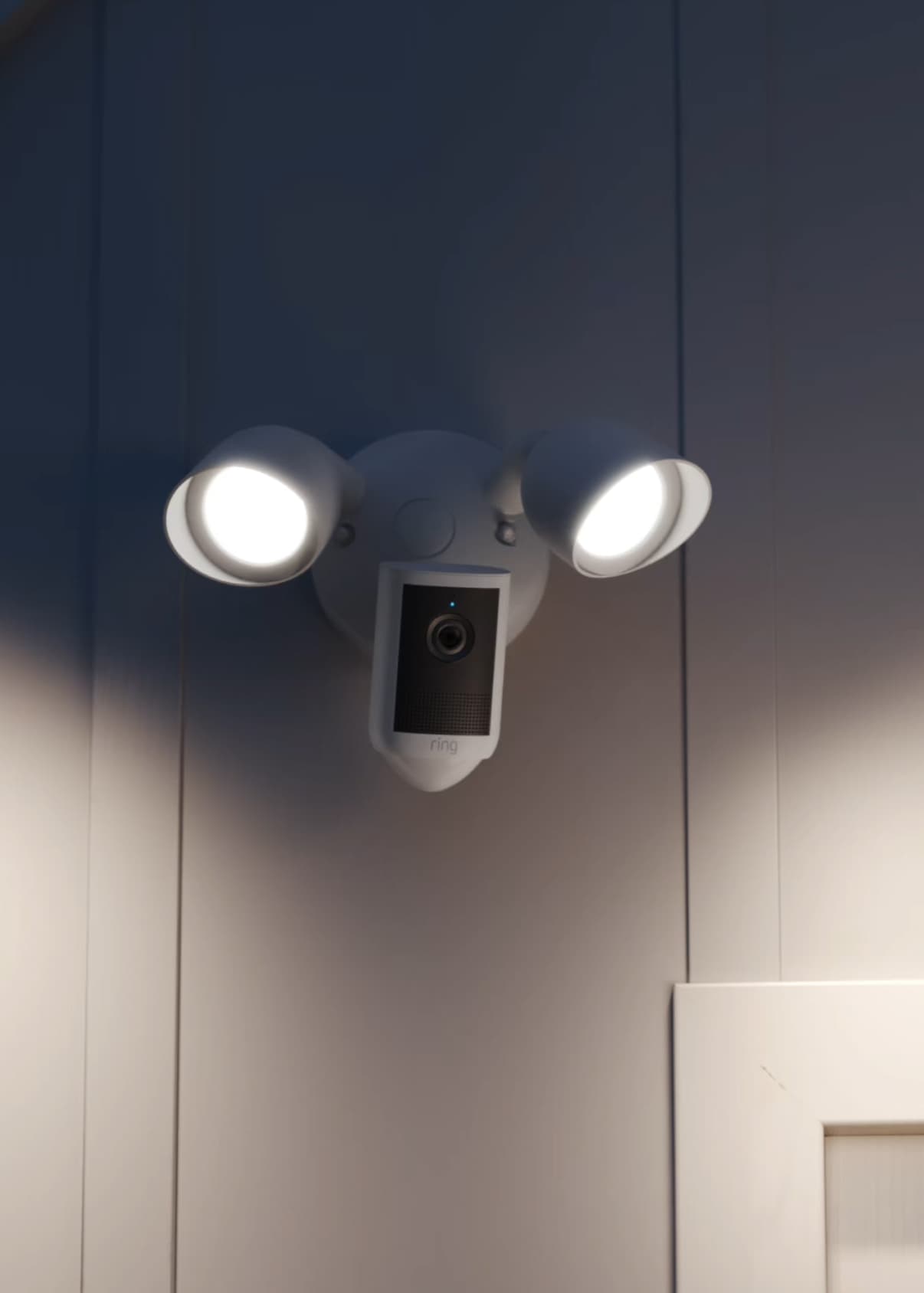 Outdoor wall with a white dual floodlight and built-in camera illuminated at dusk, casting light on the siding near a doorway.