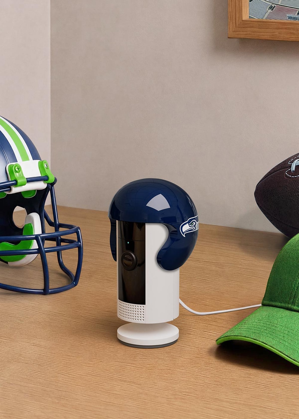 Football-themed room scene: a small indoor camera with a helmet cover on a desk beside a football helmet, ball, and green cap.