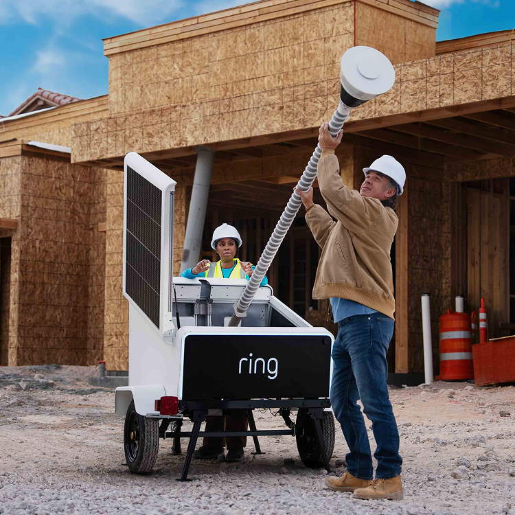 Two workers in hard hats assemble a Ring mobile security tower unit at a construction site, lifting a tall mast beside a trailer with solar panels.
