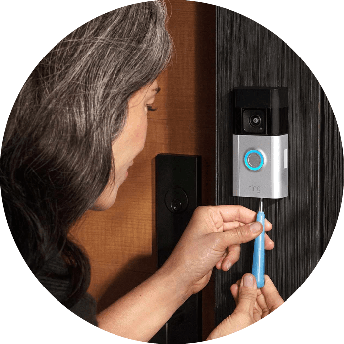 Woman securing Ring Battery Doorbell Pro with blue-lit button to wooden door frame using a screwdriver.