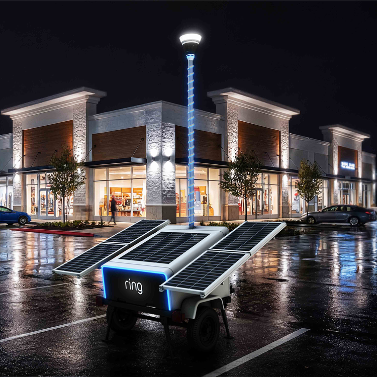 Rainy night scene at a shopping center with lit storefronts, parked cars, and a Ring Security tower with solar panels and a glowing Ring logo.