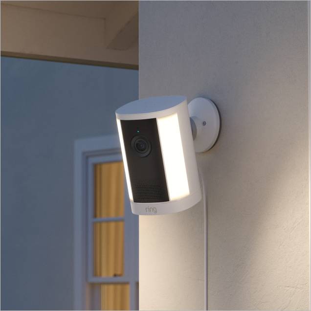 Wall-mounted outdoor security camera with dual LED lights glowing at dusk, attached to a stucco exterior near a window.