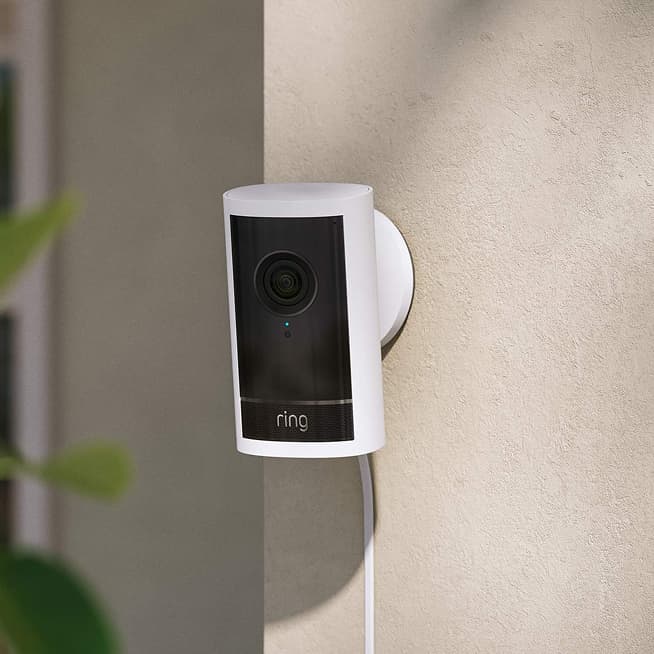 White Ring security camera mounted on a beige exterior wall, plugged in with a visible power cord, greenery blurred in foreground.