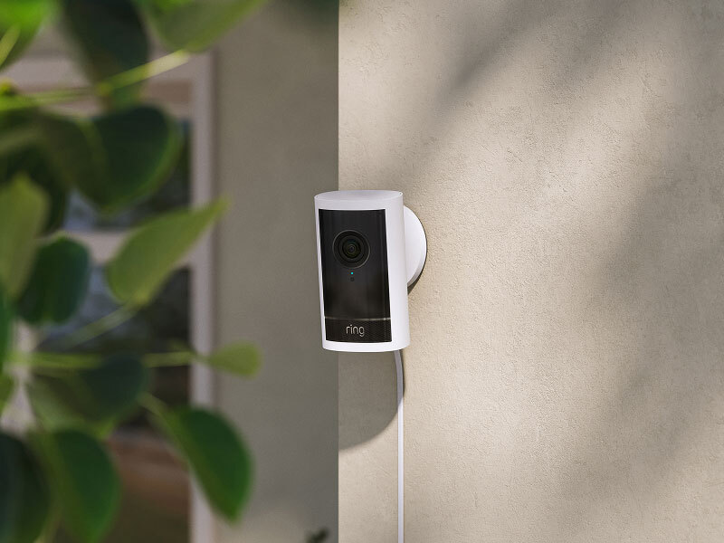 White Ring security camera mounted on a beige exterior wall, powered by a visible cord, with lens and status light facing outward.