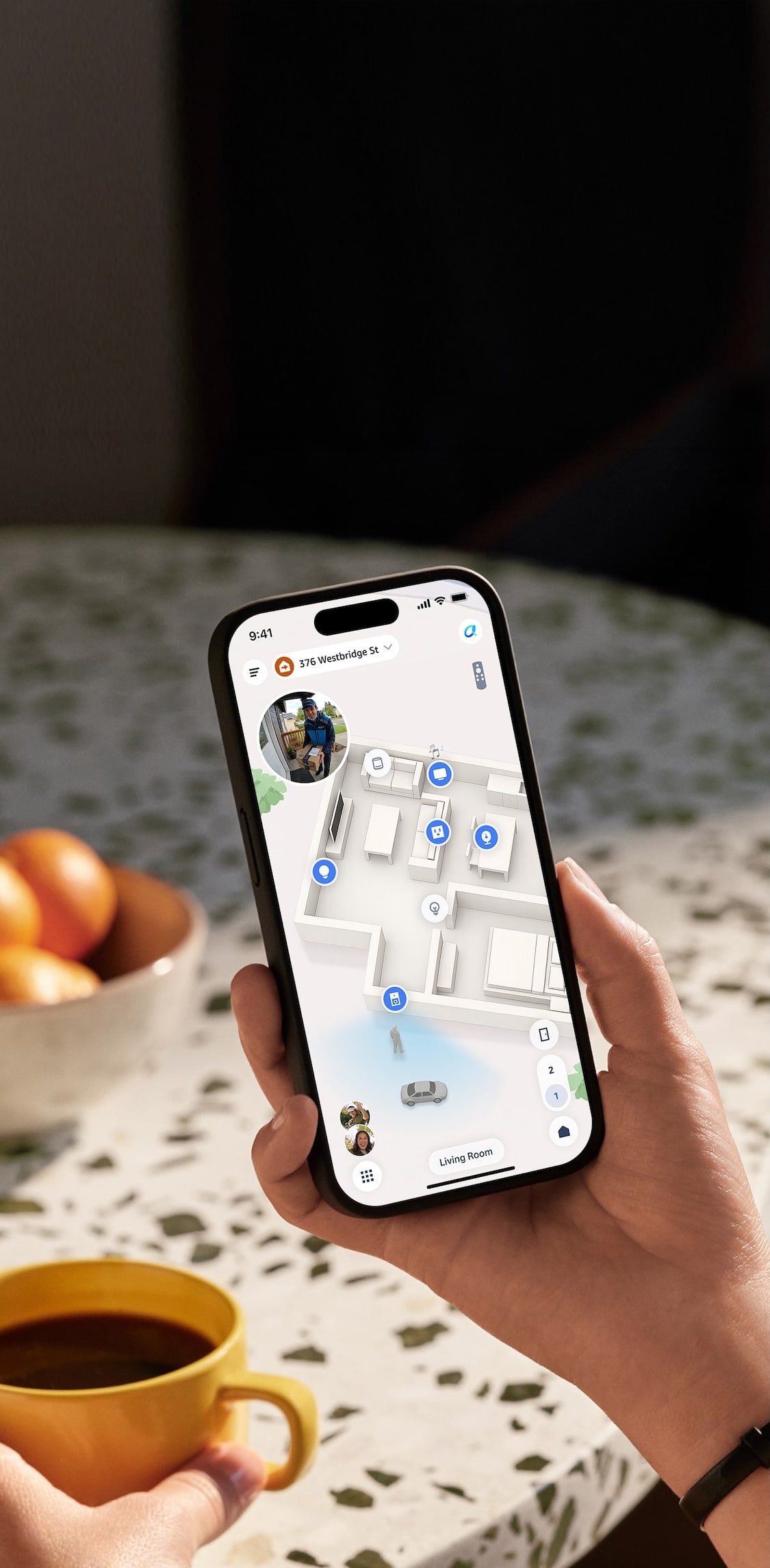 Hand holding a smartphone showing a 3D home floor plan with blue device icons, on a speckled table with a bowl of oranges nearby.