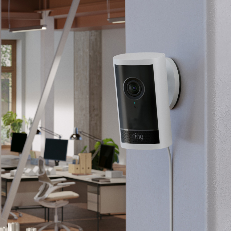 A white Ring outdoor cam pro mounted on the indoor wall of a business. Desks with computer monitors are in the background.