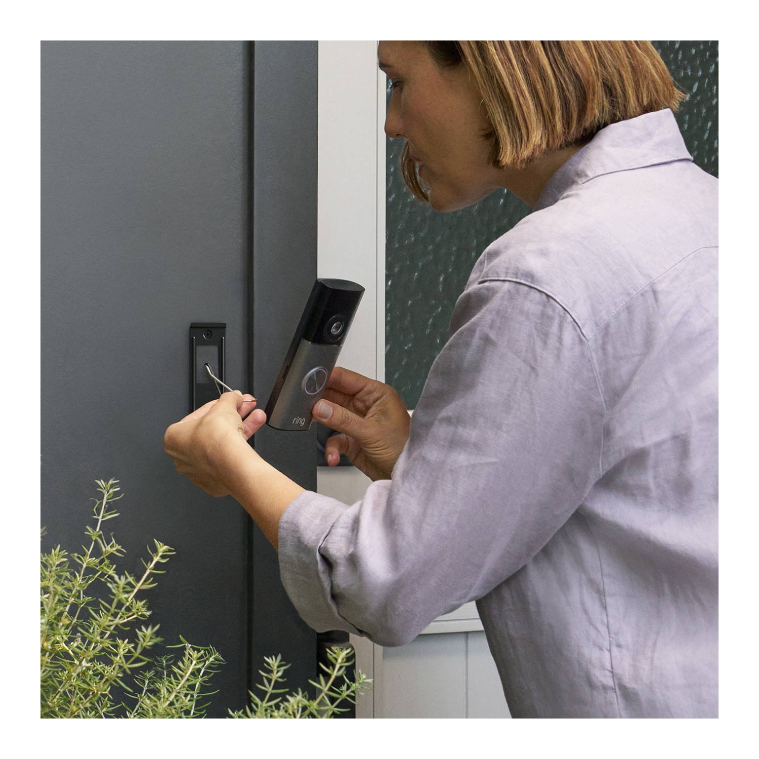 Wired Doorbell Pro (Newest Model) (Featuring Retinal 4K) - Woman installing a Wired Doorbell Pro (2nd Gen), with their hands carefully mounting the device on a wall. Part of some greenery or plants is visible in the left bottom corner of the frame.