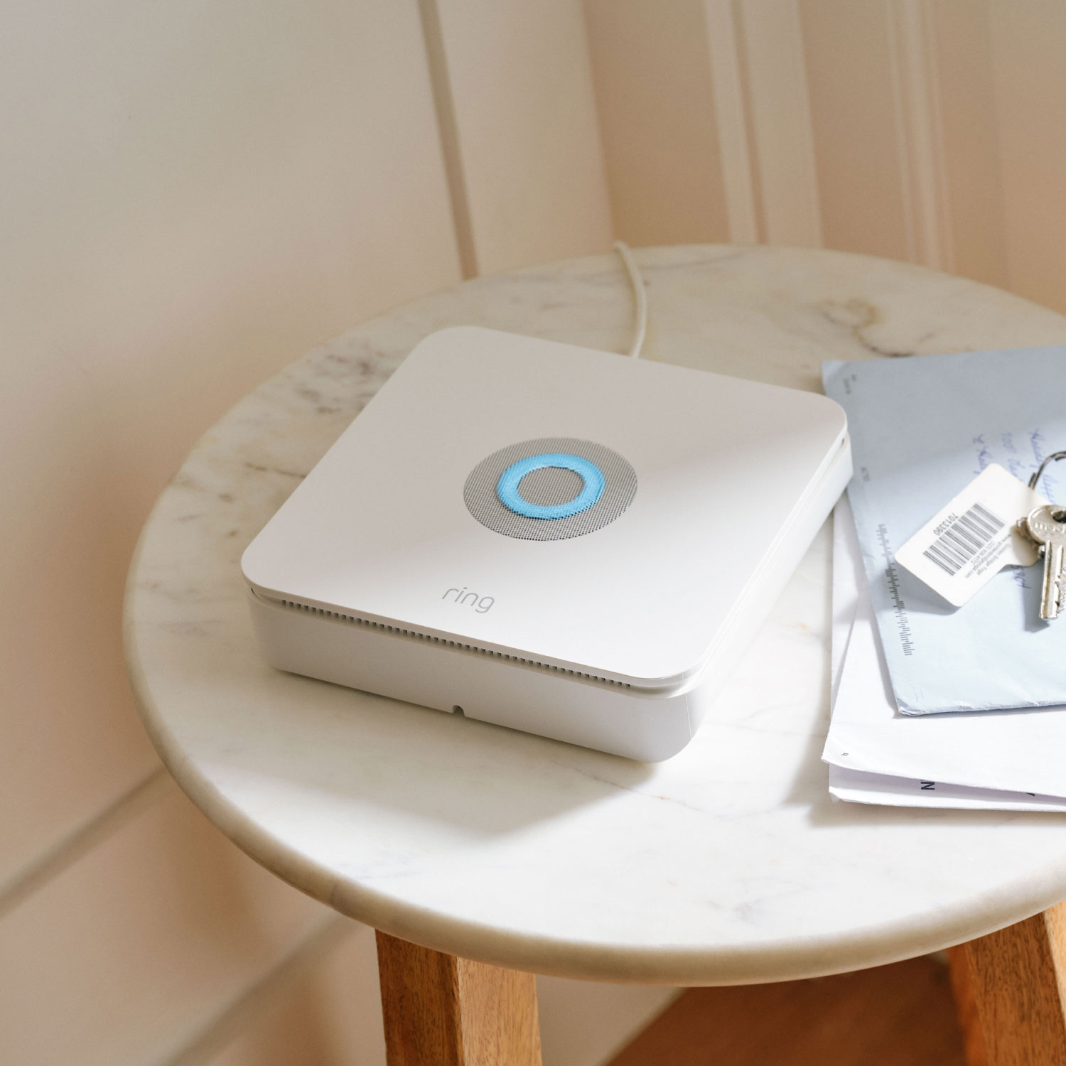 White Ring base station with a glowing blue ring on a marble side table beside mail and house keys in soft natural light.