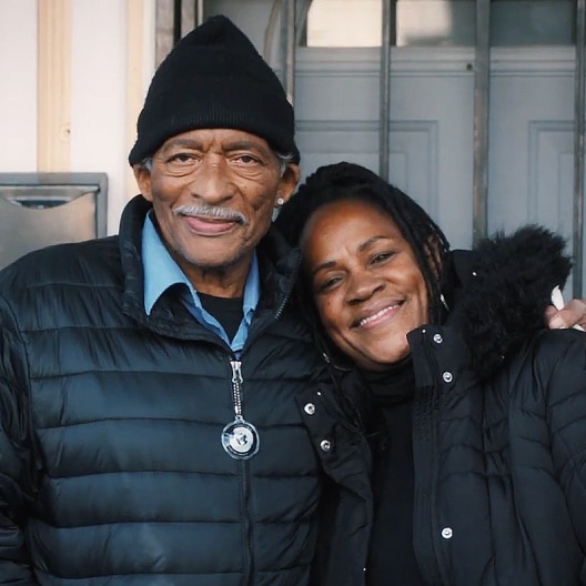 Two people smiling together outdoors, wearing black winter coats, standing in front of a light-colored building.