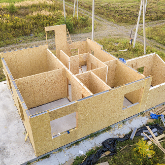 Aerial view of a small house under construction with OSB walls, open window cutouts, and framed rooms on a concrete slab in a field.