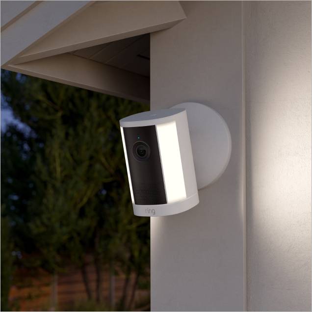 Wall-mounted Ring security camera with bright side lights glowing at night beside a storefront window and wood trim.