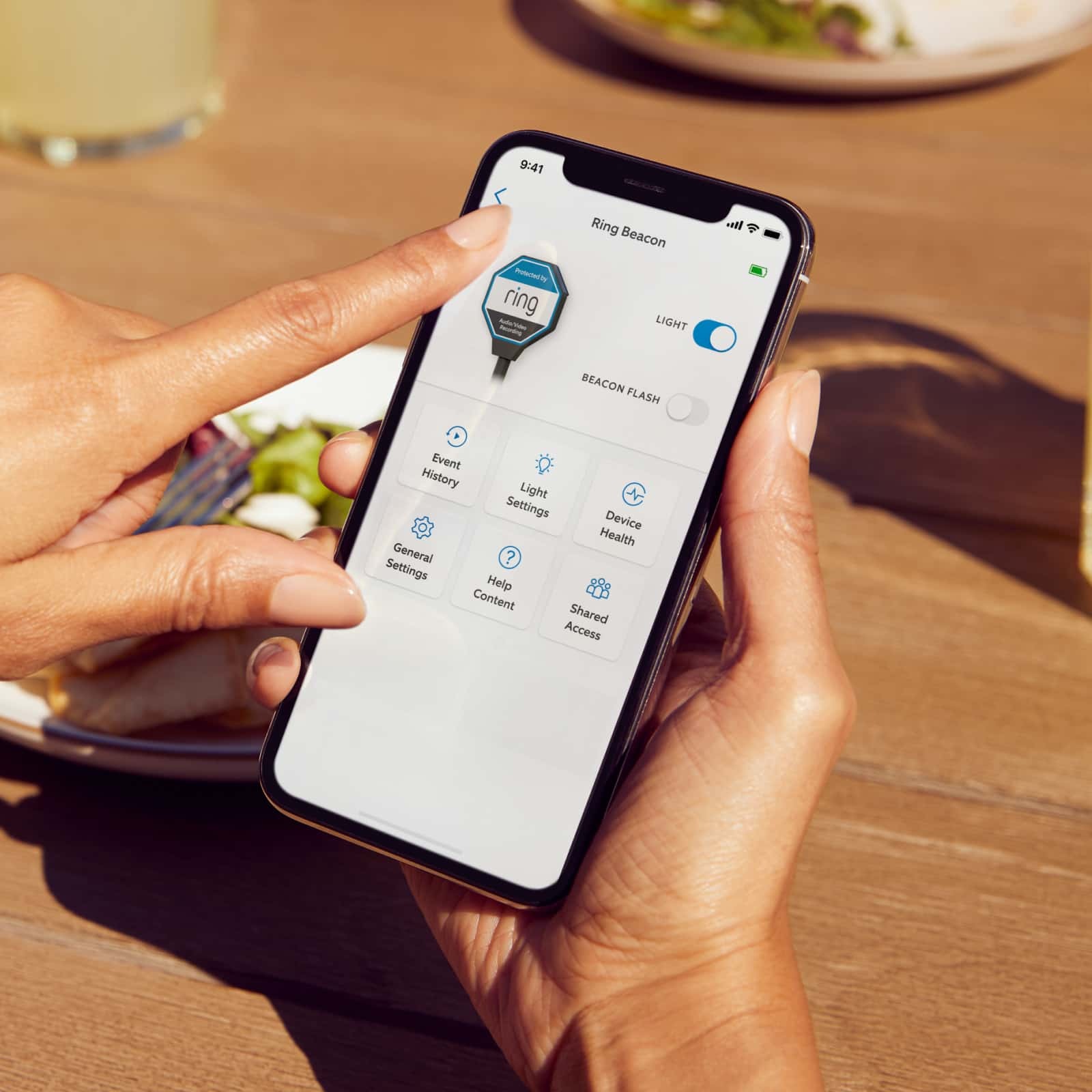 A person using the Ring app on a smartphone to control Ring Beacon light settings while sitting at an outdoor table in sunlight.
