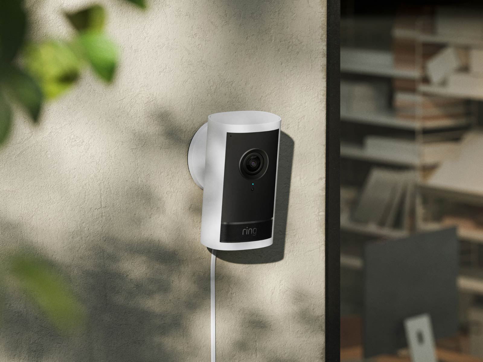 White and black Ring security camera mounted on a beige exterior wall with green foliage visible nearby.