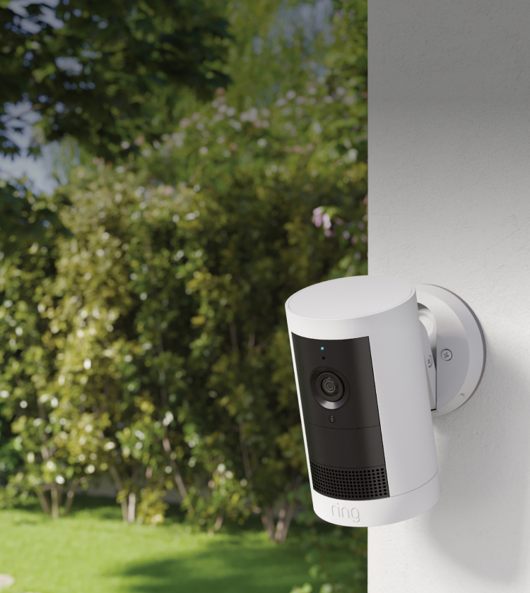 White outdoor security camera mounted on a white wall, facing a sunlit green backyard with trees and lawn.