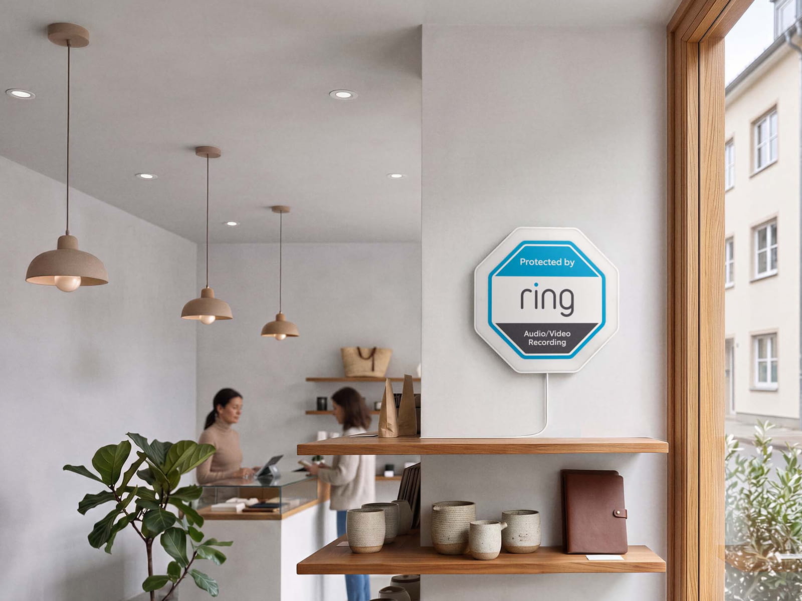Cozy shop interior with pendant lights, two people at a counter, and a wall-mounted Ring sign beside shelves and a large window.