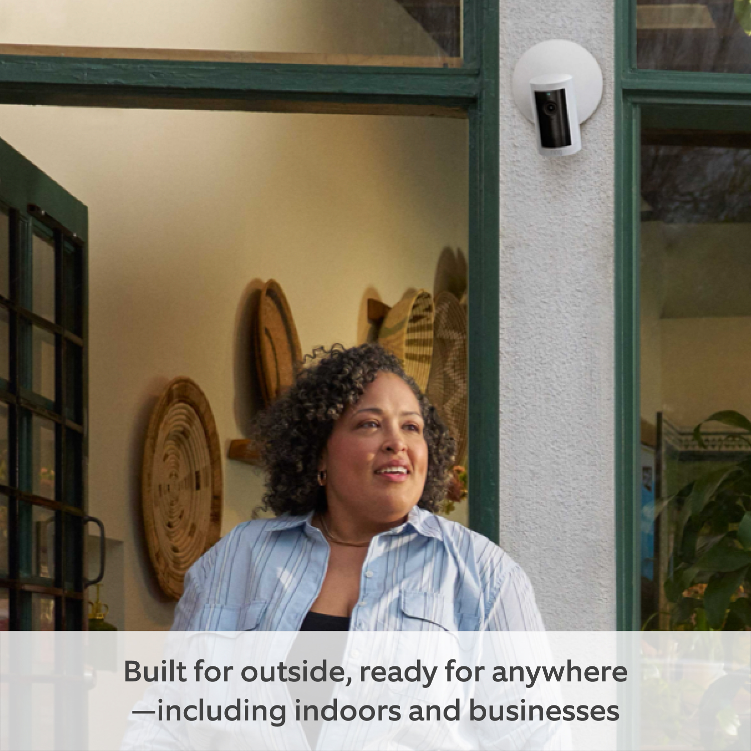 Outdoor Camera Plus (Featuring 2K Video with Ring Vision) - Power over Ethernet:Curly Hair Women standing in front of a white Outdoor Camera mounted to white wall at the corner of a green framed door.