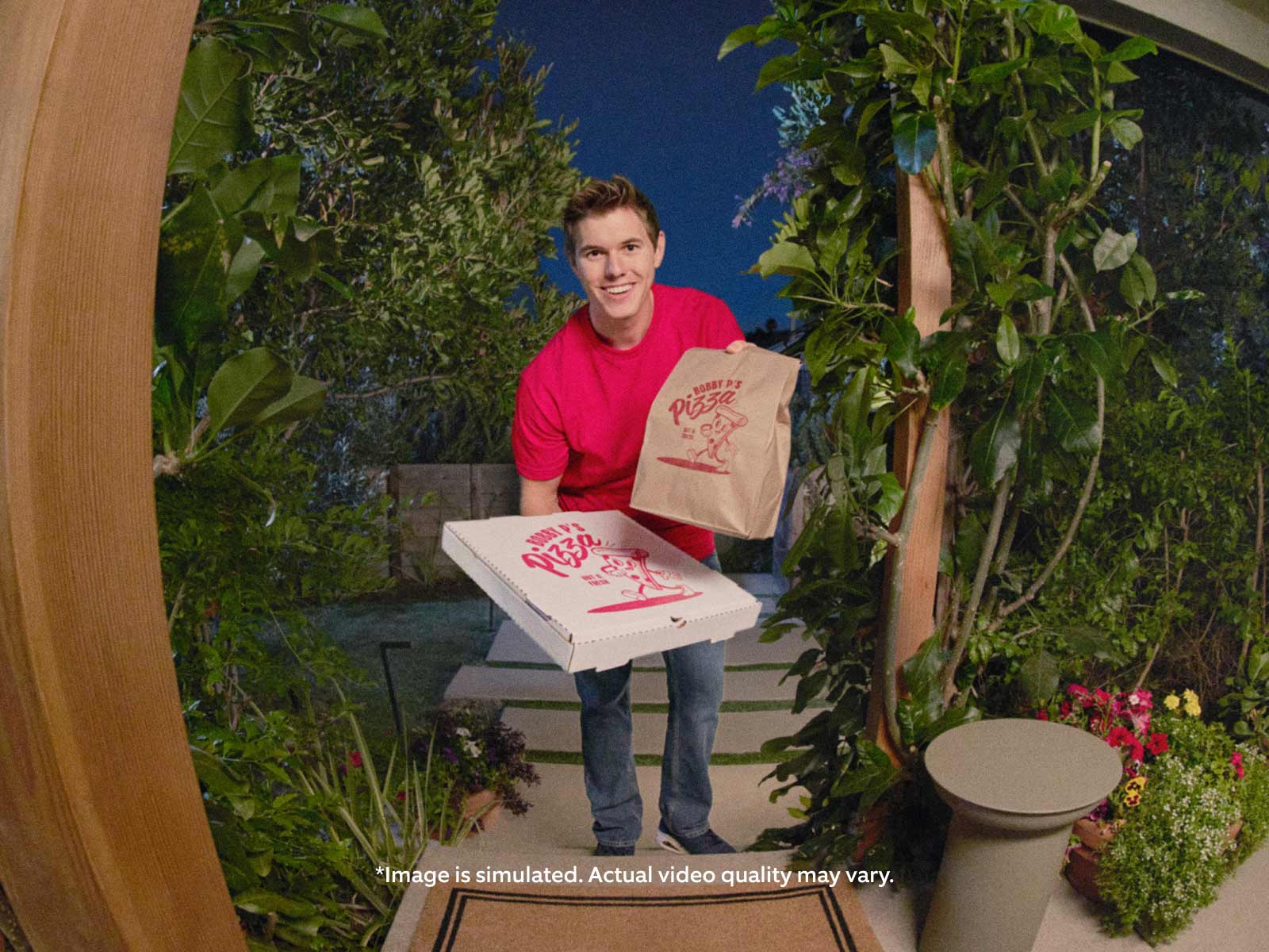 delivery person wearing a red t-shirt stands outdoors at night, holding pizza boxes and a paper bag with a company logo. They are standing at someone's front walkway It is well-lit, showing the pizza company's branding on both the boxes and paper bag.
