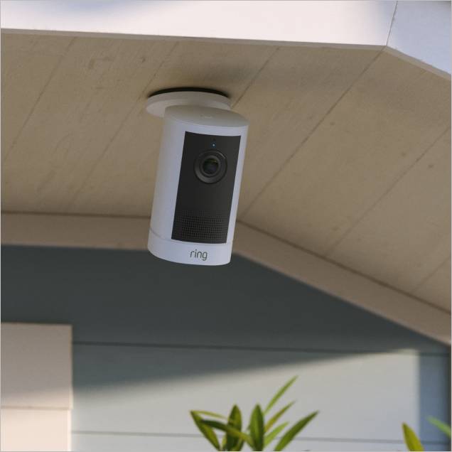 White Ring outdoor camera mounted under an eave on a house, angled downward above a doorway with plants below.