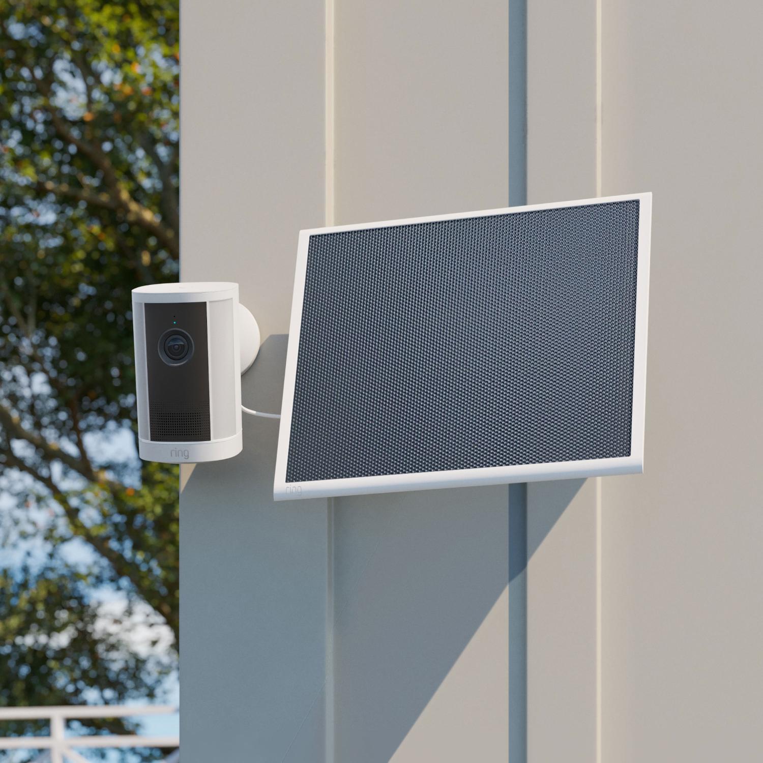 Spotlight Cam 4K Plus - Solar:Outdoor white security camera mounted on a wall, wired to a tilted solar panel, with trees and blue sky in the background.