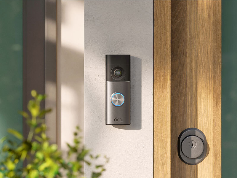 Smart video doorbell with a blue-lit button mounted beside a wooden front door and modern deadbolt, in soft daylight.