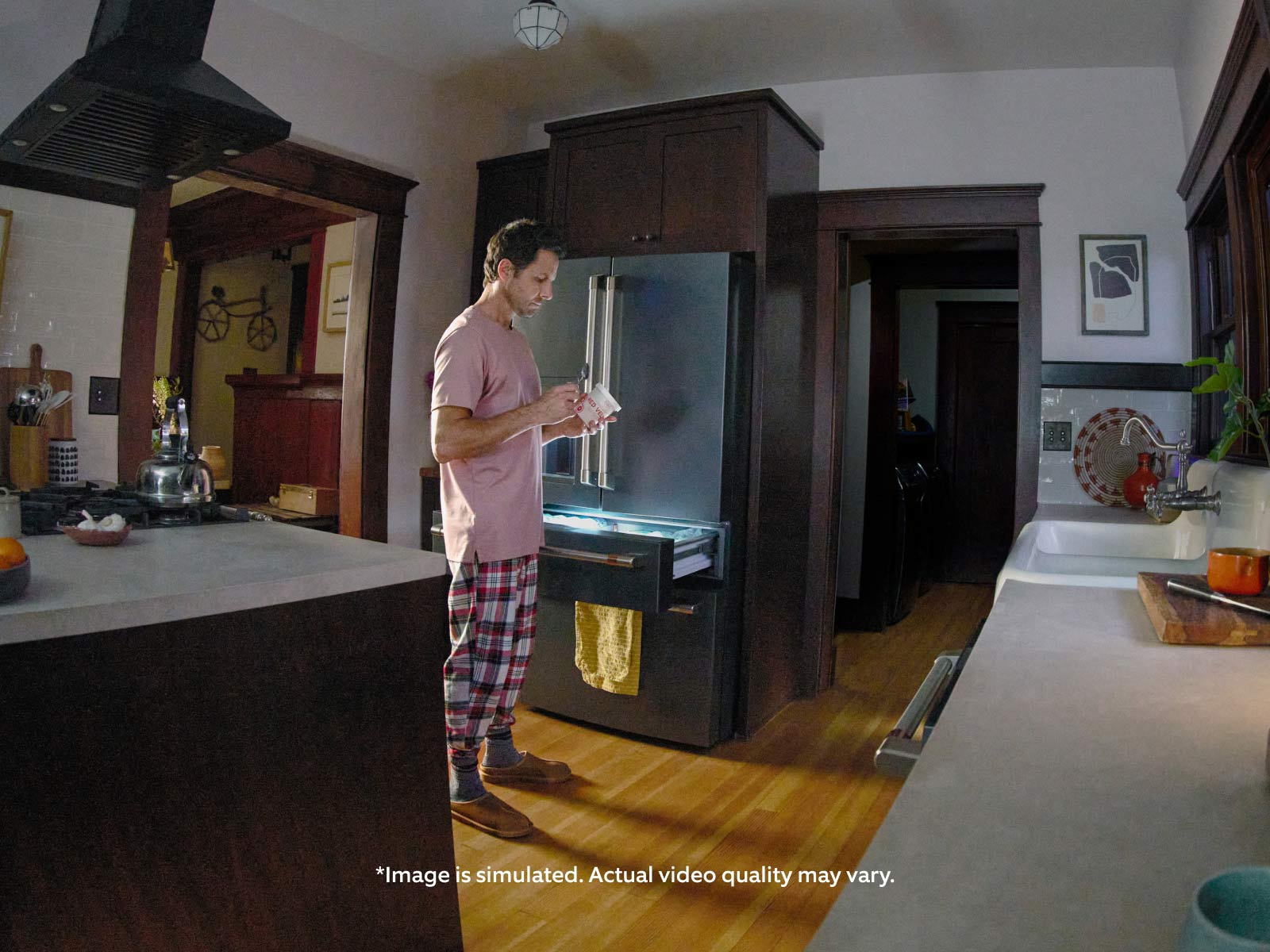 View of a home kitchen through an indoor security camera, showing a man in pajamas standing by a refrigerator eating ice cream at night. The kitchen has dark cabinets, hardwood floors, and a ceiling fan visible in the frame.
