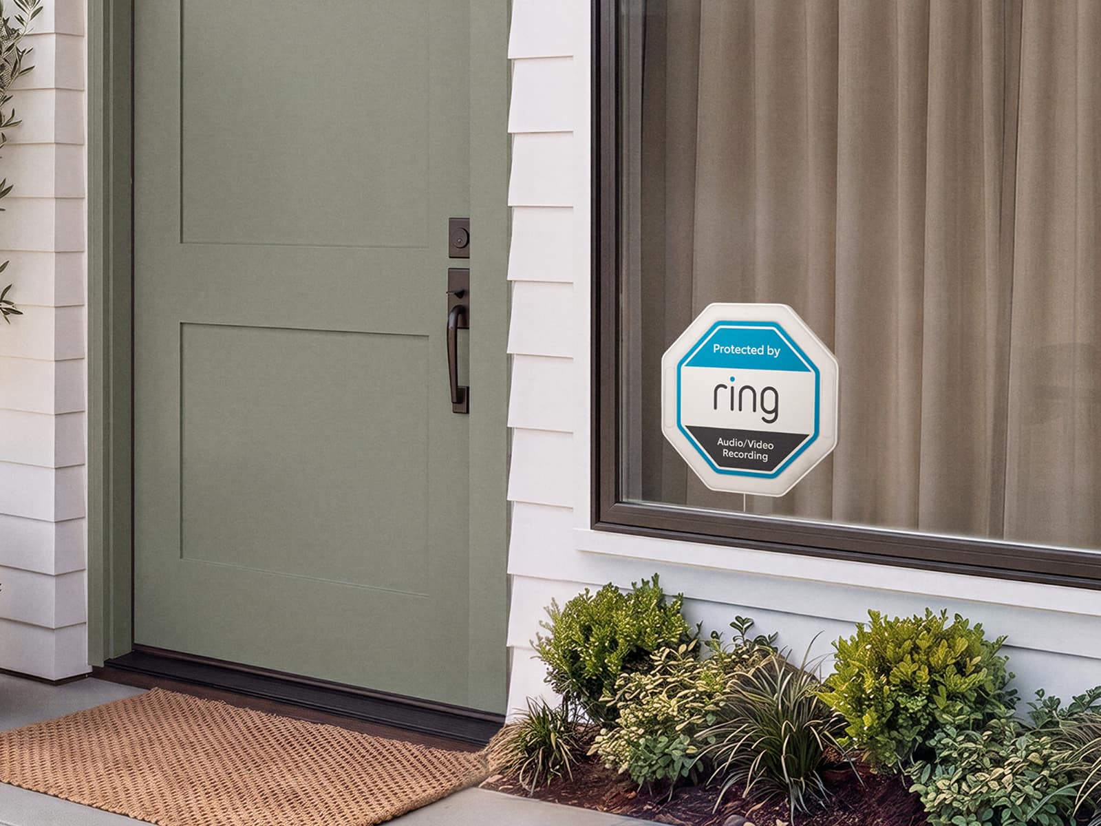 Front porch with a sage green door, doormat, and window displaying a Ring security sign above trimmed shrubs and white siding.