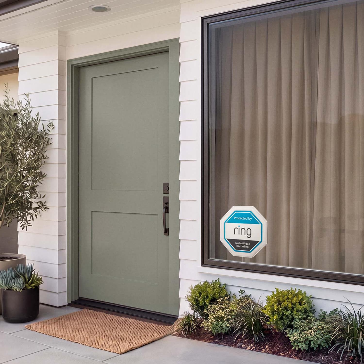Ring Indoor Illuminated Sign - Modern home entry with sage green door, welcome mat, large window with beige curtains, and a Ring security sign; potted and garden plants.