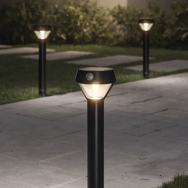 Solar-powered black path lights glowing along a stone walkway at night, with grass and shrubs in the background.