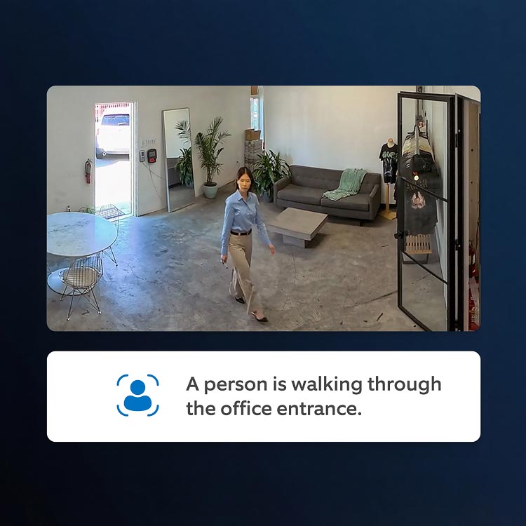 Woman in blue shirt and khaki pants walks through a bright office with concrete floor, gray sofa, plants, and a round table near an open door.