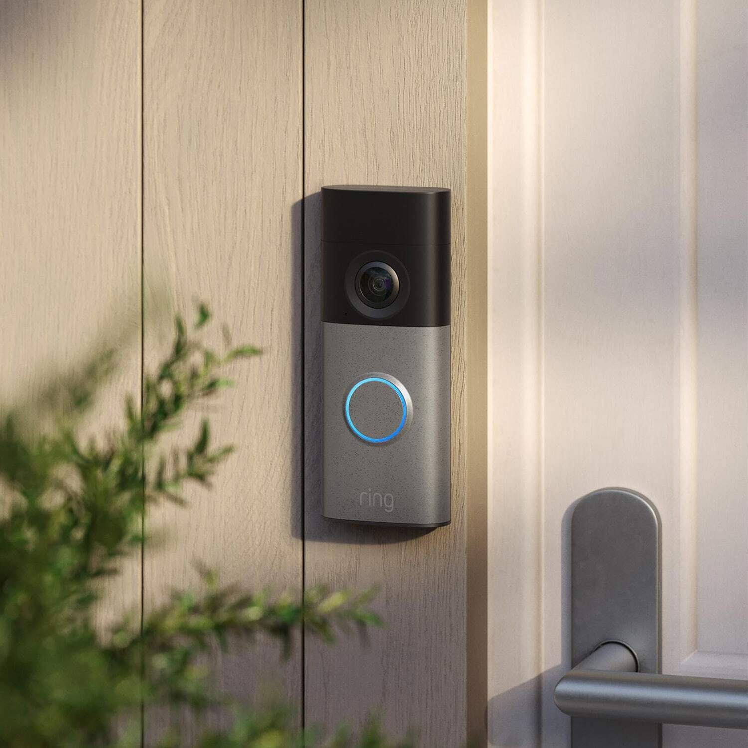 Smart doorbell with camera and glowing ring mounted on a gray front door beside a black deadbolt in soft afternoon light.