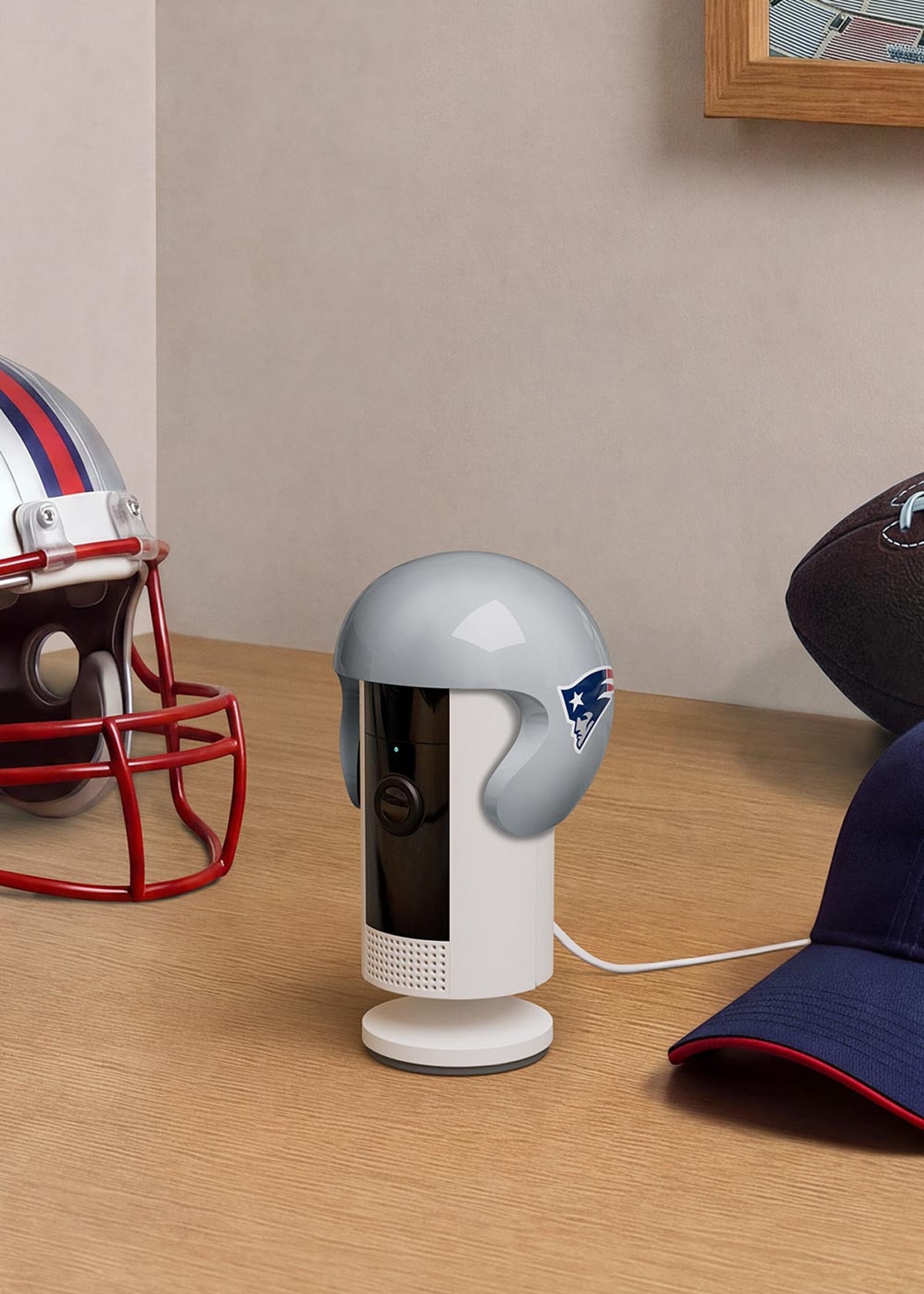 Desk scene with a small white indoor camera wearing a gray football helmet cover, surrounded by a helmet, football, and blue cap.