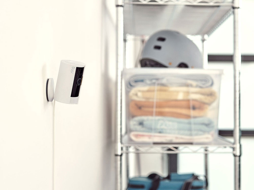 Wall-mounted indoor security camera near metal shelving with a clear bin of folded blankets and a helmet in a bright room.