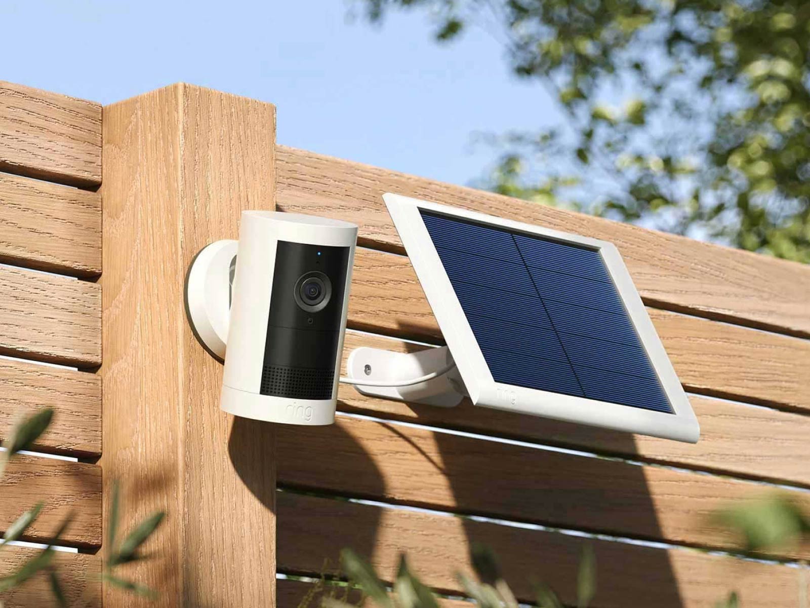 Solar-powered outdoor security camera mounted on a wooden fence, with a small solar panel beside it in sunlight.