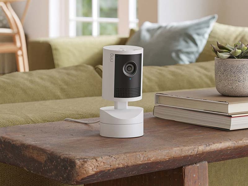 Pan-Tilt Indoor Cam 2K on a wooden coffee table beside stacked books and a potted succulent, with a green sofa behind.