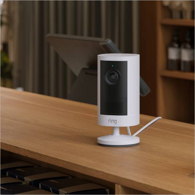 White Ring camera on a wooden counter with a tablet in the background, power cable trailing to the right.