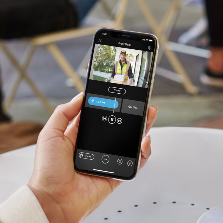 Hand holding a smartphone, the screen shows Ring app with video of a person delivering packages to the front door.