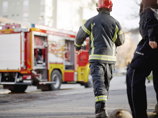 A firefighter in protective gear and a red helmet walks toward a fire truck on a city street, while another emergency responder is partially visible nearby.