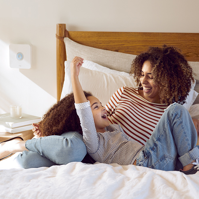 Smiling mother and daughter relax on bed in sunlit room with Ring security device on wall in background.