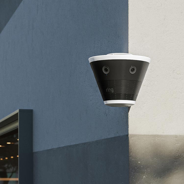 Cone-shaped black and white Ring outdoor camera with dual lenses mounted on a blue and beige building corner.