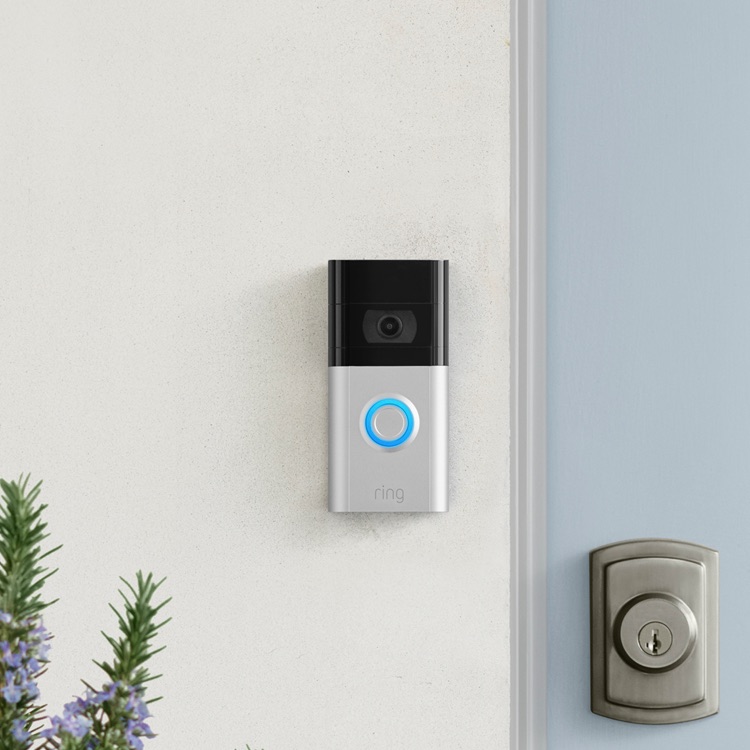 A Ring Video Doorbell with an illuminated blue ring mounted on a white wall beside a light blue door with deadbolt lock, with green plants in view.