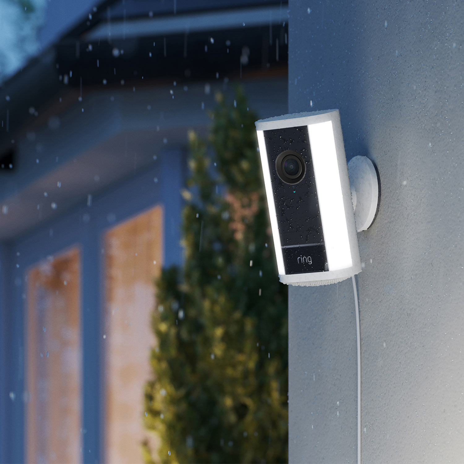 Spotlight Cam Pro (Newest Model) (Featuring Retinal 4K) - Plug-In:White Ring Spotlight Cam Pro (2nd Gen) attached to a wall at dusk in the rain with built-in LED spotlights illuminated.