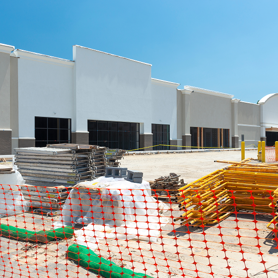 Commercial building under construction with white exterior walls, orange safety fencing, and building materials in foreground.