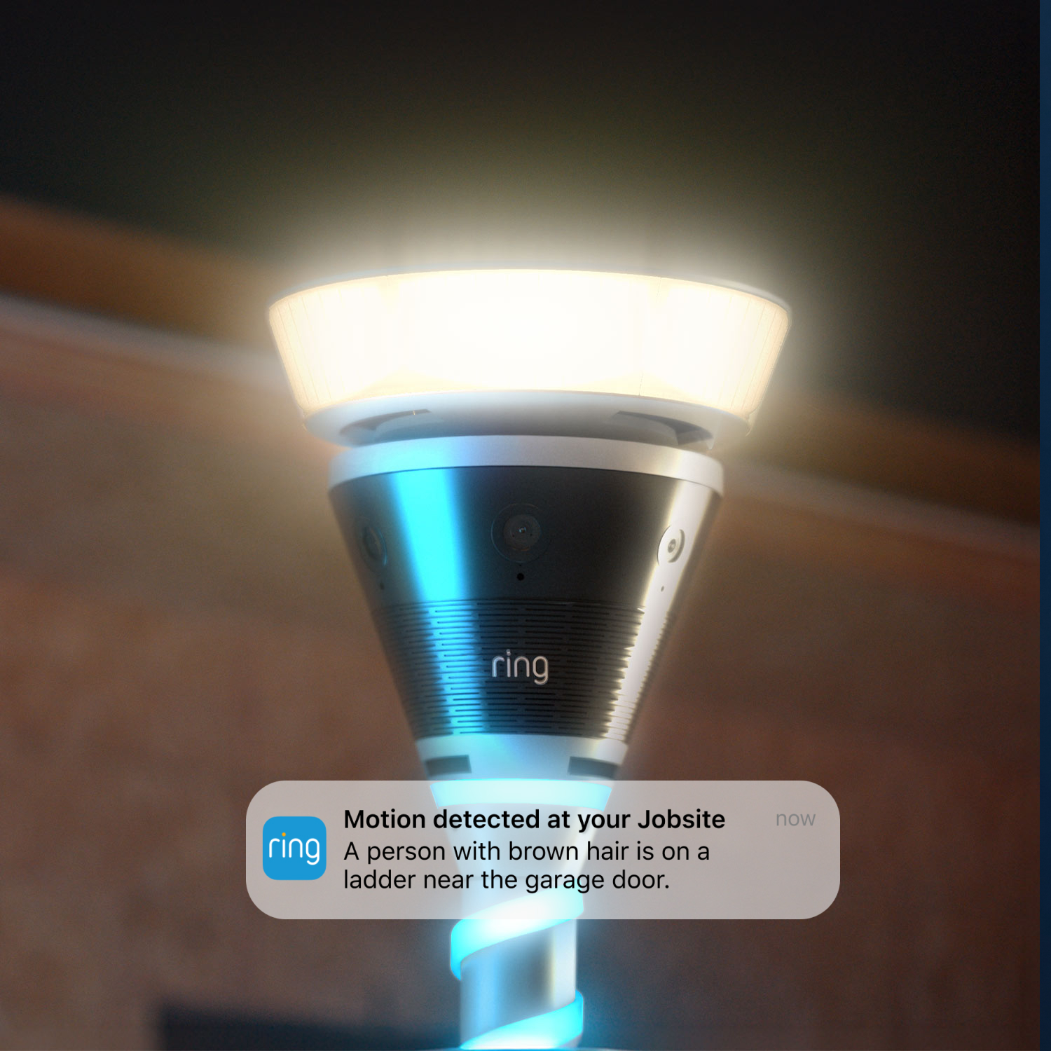 Ring Mobile Security Tower - Solar Panel Kit + 1x Battery - Close-up of a Ring pole-mounted light and camera glowing, with a phone notification overlay about motion at a jobsite near a garage door.