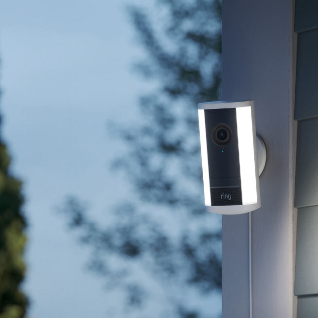 A White Ring Spotlight Cam Pro mounted to a wall at dusk illuminates suddenly. Cut to a hand holding a horizontal phone with a camera POV of the side yard where a man is seen creeping among the plants, spotlights on him.  
