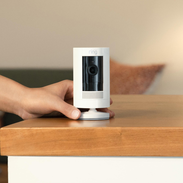 White Ring Outdoor Cam being placed on countertop next to a potted plant, with a modern living space in background.