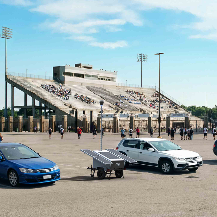 Parking lot outside a large stadium with scattered cars, people walking to the entrance, and a Ring solar-powered mobile security tower.