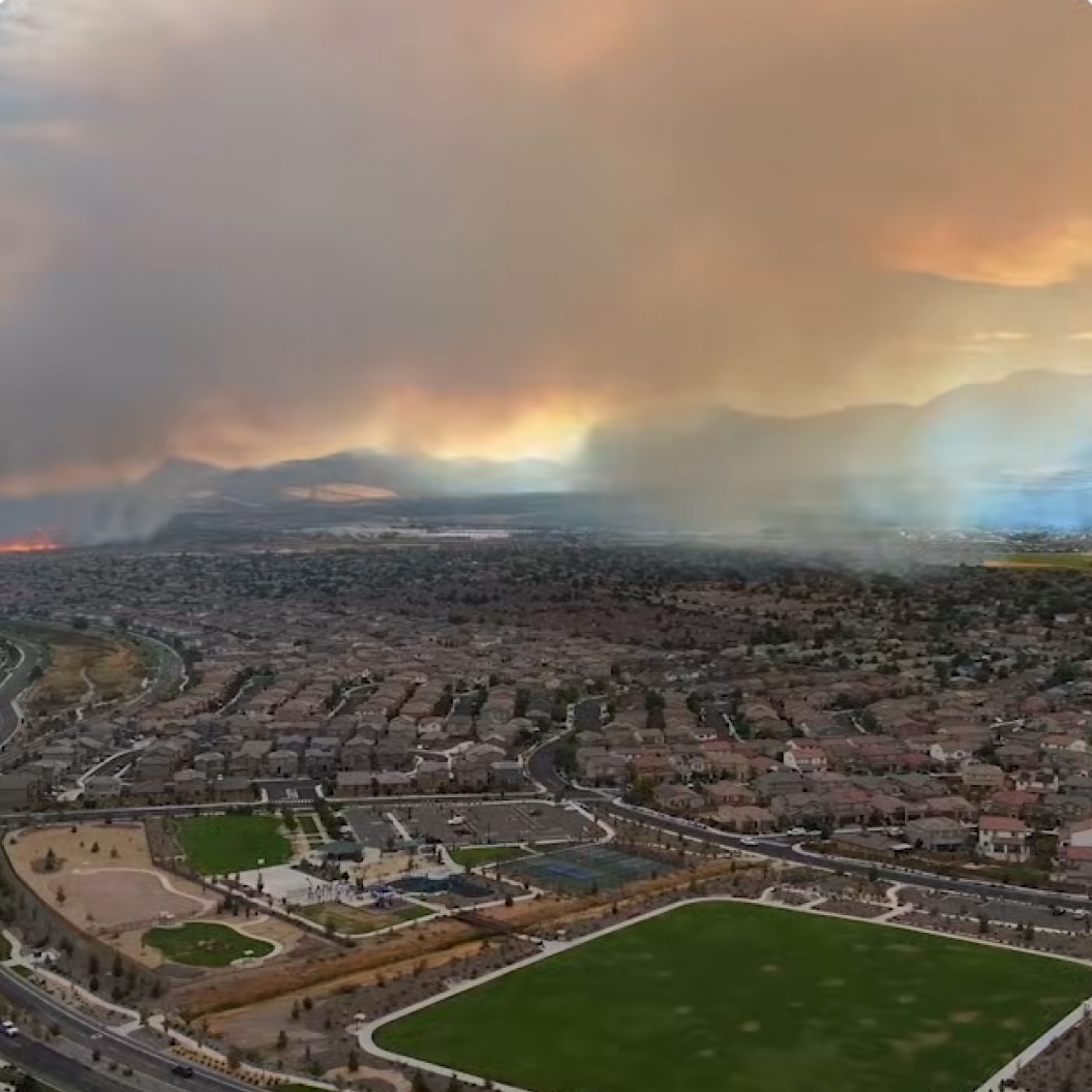 Aerial view of suburban neighborhood with smoke-filled sky from wildfire, mountains in background, and green parks below.