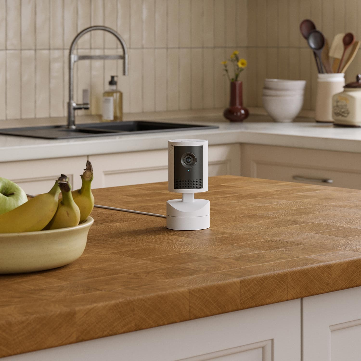 Pan-Tilt Indoor Cam 2K - Compact white indoor camera on a wooden kitchen island, with fruit bowl, sink, and utensils in the background.