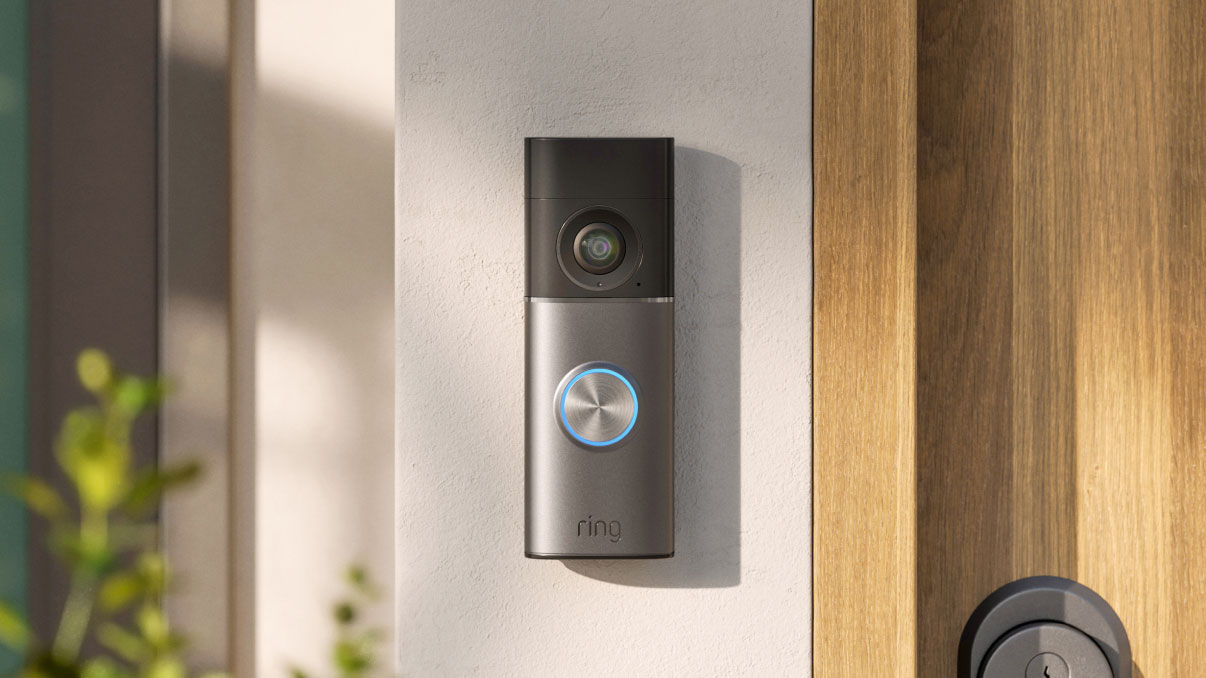 Video doorbell with camera and blue-lit button mounted on a light wall beside a wooden front door with a round deadbolt.